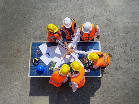 A group of six construction workers in hard hats collaborate around a table covered with plans and materials.